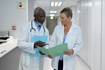 Diverse male and female doctors standing in hospital corridor looking at medical documentation - obrazy, fototapety, plakaty