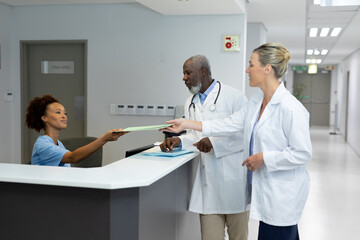 African american receptionist passing document to caucasian female doctor at hospital reception
