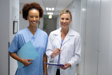 Portrait of two diverse female doctors standing in hospital corridor smiling to camera