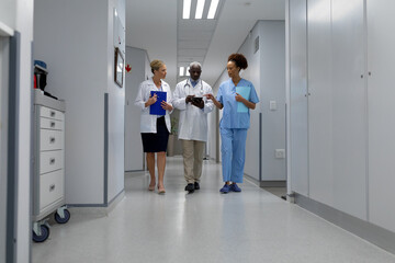 Three diverse male and female doctors walking through hospital corridor looking at digital tablet