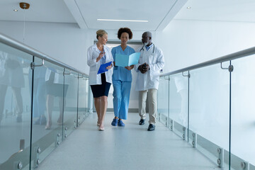 Three diverse male and female doctors walking through hospital corridor looking at document - Powered by Adobe
