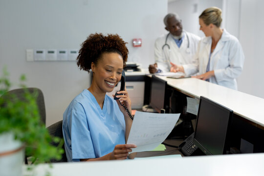 Mixed race female doctor at desk in hospital reception talking on phone and holding document