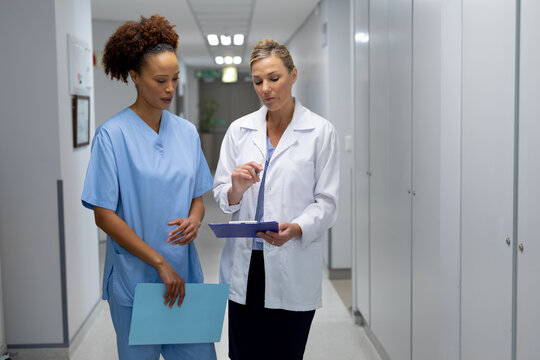 Two Diverse Female Doctors Standing In Hospital Corridor Looking At Medical Chart Document