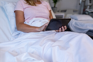 Midsection of caucasian female patient lying on hospital bed using digital tablet