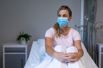 Portrait of caucasian female patient wearing mask sitting on hospital bed looking ahead