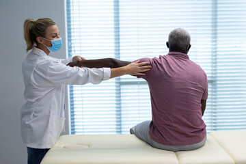 Obraz premium Caucasian female physiotherapist wearing mask stretching arm of african american male patient