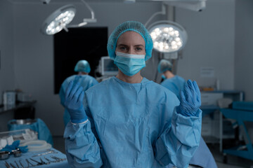 Portrait of caucasian female surgeon wearing face mask, gloves, cap and scrubs in operating theatre