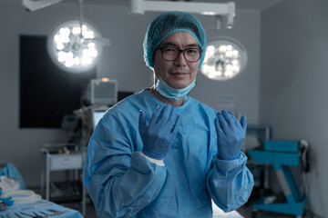 Portrait of asian male surgeon in gloves,cap and lowered face mask, smiling in operating theatre
