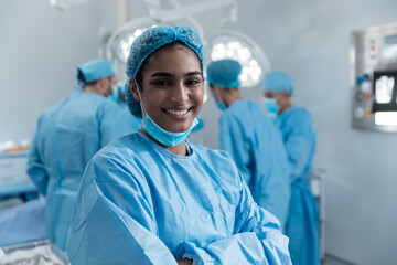 Smiling mixed race female surgeon with face mask and protective clothing in operating theatre