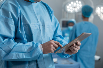 Midsection of caucasian male surgeon wearing protective clothing using tablet in operating theatre
