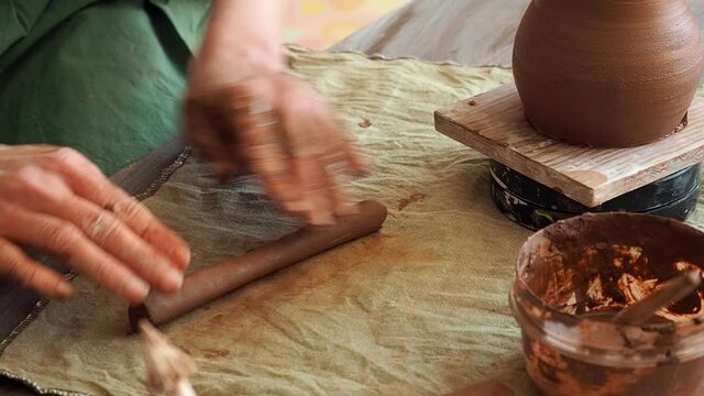 A woman potter in a green apron rolls out red clay for a potter handle