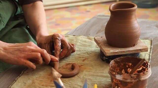 A woman potter in a green apron sculpts handle for a pot from red clay