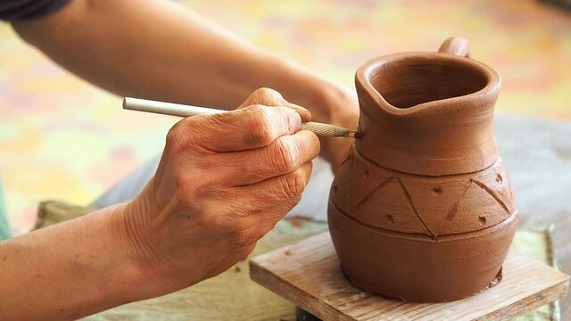 The female hands of a master potter draw a pattern on a clay pot.