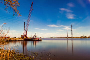 Fototapeta premium A fine art landscape image of a long exposure view of Nanticoke river on the coast of Vienna, Maryland near Chesapeake bay. A crane, a docked vintage cargo or tow boat and high voltage line tower seen