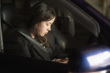 A girl with a phone in her hands and headphones in her ears is sits in the car and listening a music or audiobook