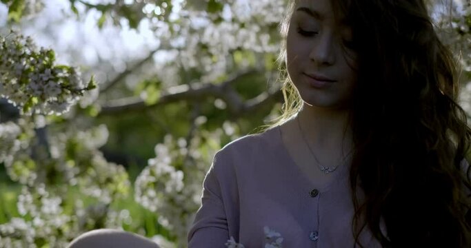 Girl with long light brown hair, grey eyes dressed in airy pink dress holds summer hat and flowering apple tree  twig. She tries the to attach the twig to hat but fails and drops it. Filmed outdoors a