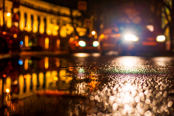 Autumn rainy night in the city. Headlights of approaching cars. Parked cars. Residential buildings in the city center. Colorful colors. Close up view from the level of the puddle on the pavement.
