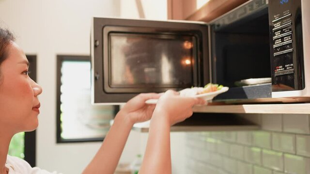 slow motion scene of asian young woman opening microwave oven to lay dish of food for defrost in the kitchen at home for comfortable lifestyle concept