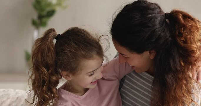 Happy young mother and little adorable cheery daughter sit on sofa cuddling laughing fooling touch noses enjoy tender moment spend time together at home. Carefree family playtime, fun and love concept