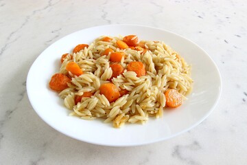 On a white marble table, cooked orzo pasta with sliced carrot and black pepper is served on a plate. Closeup view, selected focus