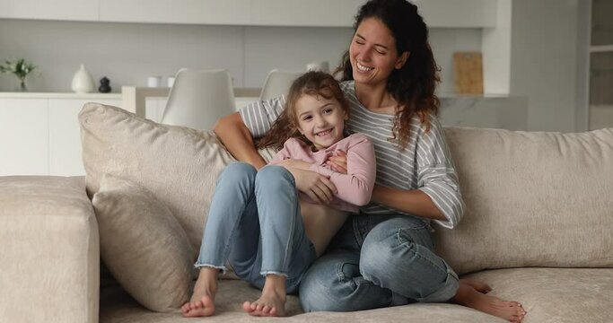 Cheerful young mother play with little daughter, tickling laughing kid girl while relax together on comfy sofa in living room. Family have fun, carefree leisure activity with children at home concept