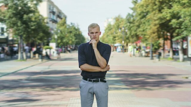 Tilt-down time lapse of attractive young businessman standing outdoors in busy street with pensive expression while people are walking around