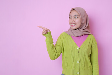 Charming pleased young Muslim Asian teenager smiling points aside on copy space shows advertisement wears muslim casual clothes against pink background