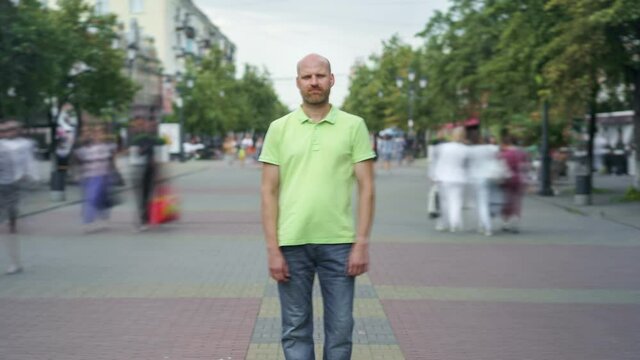 Zoom-in time lapse of serious guy in casual clothing standing in busy street among crowd of people and looking at camera. Lifestyle and people concept.