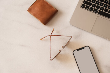 Flatlay of blank screen mobile phone, laptop, wallet, glasses on marble table. Home office desk...