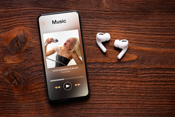 Music player on screen of mobile phone and wireless earbuds on wooden surface