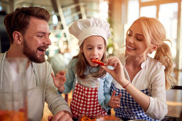 A little daughter eating paprika while preparing a meal at home with her parents. Family, together, home