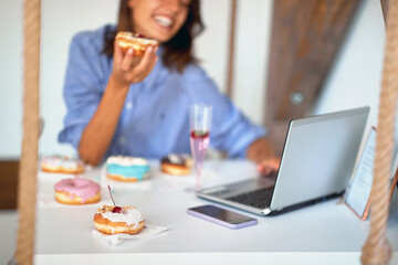 A woman is having a good time while eating delicious donuts and using a laptop in a pastry shop. Pastry shop, dessert, sweet