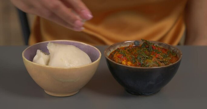 Two bowls on table with West African traditional dishes. Unrecognizable woman eating thick soup and white sticky dough with fingers. Fufu. Ogbono. Common local food. Nigerian cuisine