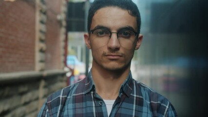 Slow motion close-up portrait of good-looking young Arab man wearing glasses looking at camera with serious face standing outside. People and emotions concept. - Powered by Adobe