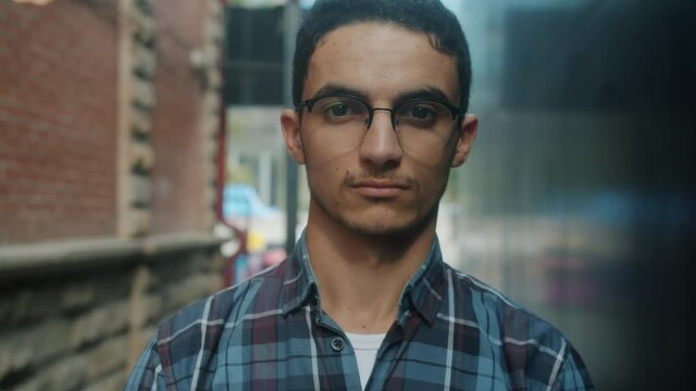 Slow motion close-up portrait of good-looking young Arab man wearing glasses looking at camera with serious face standing outside. People and emotions concept.