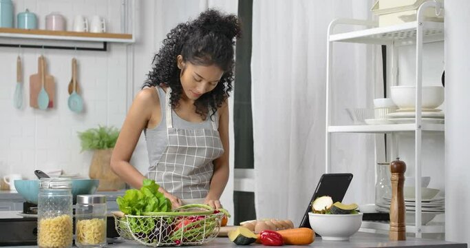 A young, happy and beautiful Asian woman with curly long hair wearing grey gingham apron. She slide vegetable on chopping block with knife and looking at the tablet for food recipe in modern kitchen.