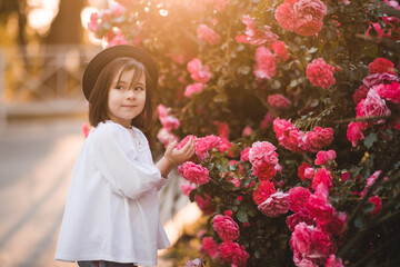 Cute stylish child girl 5-6 year old wear trendy clothes and hat posing over flower rose outdoors in park. Having fun. Childhood. Happiness.