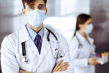 Young male doctor standing with arms crossed in clinic. Smart physician wearing medical protective mask in modern hospital. Medicine and Corona virus concept