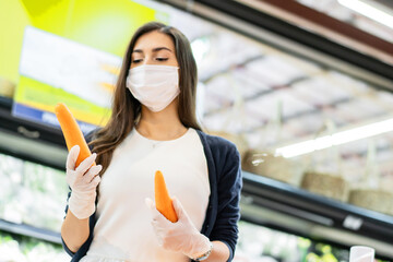 Woman wearing protective face mask and rubber glove holding carrot at vegetable grocery department store. shopping at supermarket in new normal lifestyle concept during Coronavirus pandemic