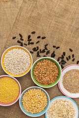 Portions of various cereals - wheat, peas, buckwheat, millet, oatmeal, barley in a colored dish against a background of burlap and scattered sunflower seeds, flat lay, copy space