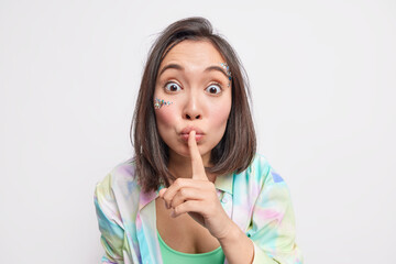 Fototapeta premium Hey hear my secret. Surprised brunette Asian woman thrilled by great news makes shush gesture dressed in casual clothes isolated over white background tells rumors has mysterious expression.