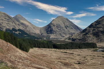Mountains of Glen Etive in Scottish Highlands on a sunny day with blue sky and light cloud. Valley and forest in front of mountains. House from James Bond Skyfall can be seen in far distance.