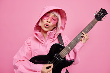 Carefree female singer enjoys music sings favorite song plays electrical guitar dressed in anorak...