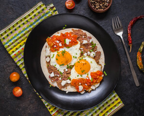 Huevos rancheros, Mexican fried egg on a wheat tortilla with tomato salsa, bean paste and feta cheese on a black plate on a light concrete background.