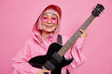 Obraz premium Photo of pleased fashionable female musician plays electric guitar smiles pleasantly wears heart shaped sunglasses anorak with hood poses against pink background. Monochrome shot. Music concept