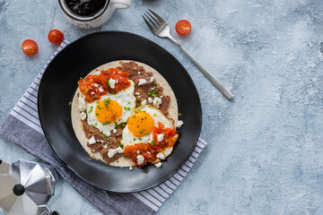 Huevos rancheros, Mexican fried egg on a wheat tortilla with tomato salsa, bean paste and feta cheese on a black plate on a light concrete background.