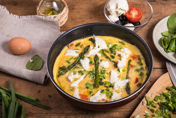 Cooking omelet with asparagus, spinach and tomatoes in a pan, homemade rustic breakfast. Wooden table with ingredients and frying pan with frittata
