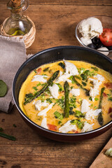Cooking omelet with asparagus, spinach and tomatoes in a pan, homemade rustic breakfast. Wooden table with ingredients and frying pan with frittata