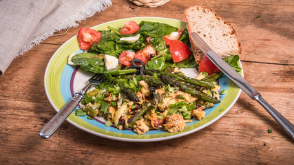 American fried scrambled eggs with asparagus, spinach and tomatoes in a large plate on a rustic wooden table - a simple rustic breakfast