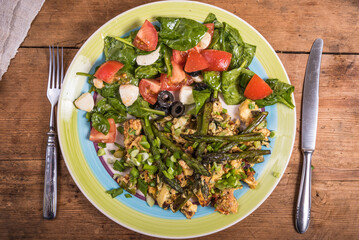 Fried scrambled eggs with asparagus, spinach and tomatoes in a large plate on a rustic wooden table, top view, close-up - simple rustic breakfast